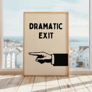 May include: A framed print featuring the words "DRAMATIC EXIT" in black, with a pointing hand illustration. The frame is light brown, and the background is a light beige colour. The print is displayed against a window with a coastal view.