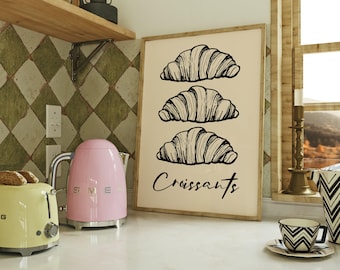 May include: A framed print featuring three croissant illustrations with the word "Croissants" in a script font. The print is displayed on a kitchen counter with a pink kettle, a yellow toaster, and a patterned tile backsplash.