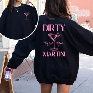 Puede incluir: Sudadera negra con texto rosa que dice "DIRTY Social Club EST 1952 MARTINI" y un gráfico de copa de martini rosa.