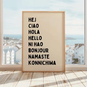 Puede incluir: Un cuadro con un fondo beige y texto negro que incluye saludos en varios idiomas: HEJ, CIAO, HOLA, HELLO, NI HAO, BONJOUR, NAMASTE y KONNICHIWA. El marco es de color marrón claro y la impresión se muestra cerca de una ventana.