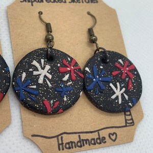 Puede incluir: Un par de pendientes redondos negros con un acabado brillante. Los pendientes presentan diseños de fuegos artificiales rojos, blancos y azules. Los pendientes están hechos a mano y tienen un cierre de gancho.