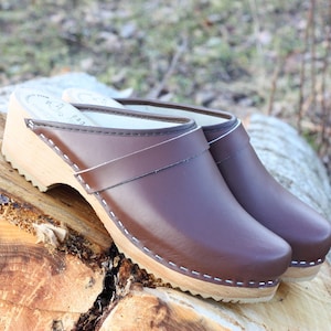 Puede incluir: Un par de zuecos de cuero marrón con suelas de madera. Los zuecos tienen un diseño clásico con punta redondeada y una correa en la parte superior. Las suelas son de madera clara y miden aproximadamente 5 cm de alto. Los zuecos están sobre un tronco de árbol.