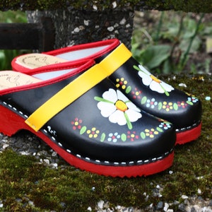 May include: A pair of black clogs with a red sole and heel. The clogs feature a yellow strap and are decorated with a white daisy and colorful floral accents. The clogs are approximately 10 inches long.