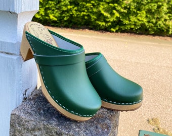 May include: Green leather clogs with wooden soles and heels. The clogs have a rounded toe and a strap across the top. White stitching accents the edges of the green leather. The clogs are displayed outdoors.