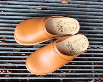 May include: A pair of light brown leather clogs with a rounded toe and wooden sole. The clogs have a stitched seam around the edge and the text "SKANE TOFFELN" is visible on the insole.