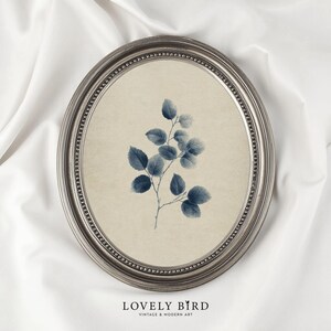 May include: An oval framed print featuring a botanical illustration of a leafy blue branch against a beige background. The frame is silver with a beaded edge. The text "LOVELY BIRD" is at the bottom.