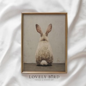Könnte beinhalten: Ein gerahmter Vintage-Print mit einem Hasen von hinten. Der Hase hat hellbraune Ohren und einen flauschigen weißen Körper. Der Druck befindet sich in einem goldenen Rahmen, mit dem Text "LOVELY BIRD" und "VINTAGE & MODERN ART" unten.