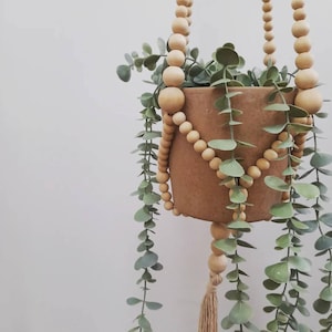 May include: A hanging planter with a terracotta pot and faux eucalyptus. The planter is suspended by a beaded wooden chain. A tassel hangs from the bottom of the pot. The leaves are a muted green colour.