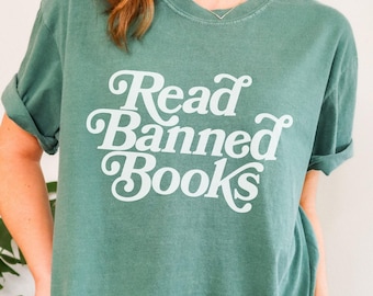 May include: Light green t-shirt with the text "Read Banned Books" in white script. The shirt has a relaxed fit and rolled-up sleeves. The bottom right corner of the image has a light orange banner that says "Light Green".