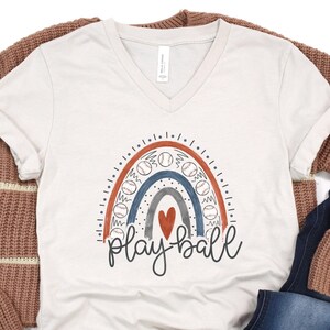 May include: A cream-colored V-neck t-shirt featuring a baseball-themed rainbow design with the words "play ball" in a script font. The rainbow includes baseballs and a red heart. The shirt is paired with a brown sweater and blue jeans.
