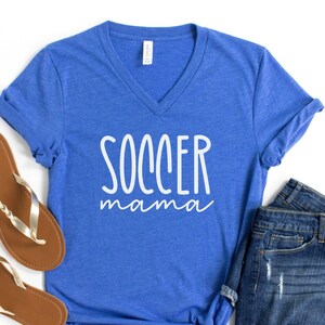 May include: A royal heather blue V-neck t-shirt with the white text SOCCER mama printed on the front, paired with a pair of brown flip-flops and dark blue denim shorts. The shirt features short sleeves and a casual, relaxed fit.