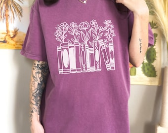 May include: Berry-colored t-shirt with a white graphic of books with flowers. The shirt has short sleeves and a crew neck. The word "Berry" is printed in white on an orange rectangle at the bottom of the image.