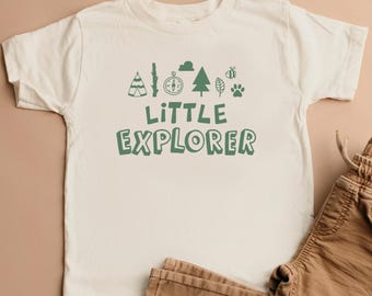 May include: Cream-colored t-shirt with the text "LITTLE EXPLORER" in green. The shirt features green graphic icons of a tent, compass, tree, bee, leaf, and paw print. Brown pants are visible in the bottom right corner.