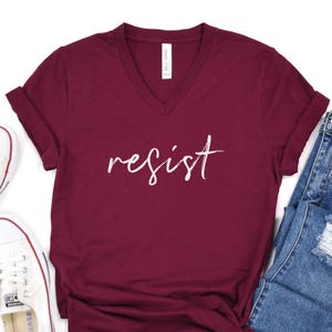 May include: A burgundy v-neck t-shirt with the word "resist" in white script. The shirt is folded, with a pair of blue jeans and a white sneaker visible in the background. The shirt is made of a soft material.