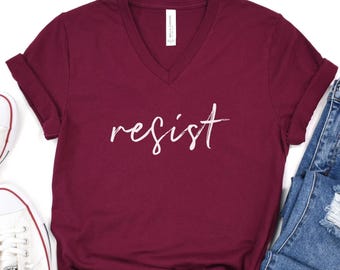 May include: A burgundy v-neck t-shirt with the word "resist" in white script. The shirt is folded, with a pair of blue jeans and a white sneaker visible in the background. The shirt is made of a soft material.