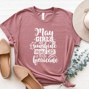 May include: Dusty rose t-shirt with the text "May Girls are sunshine mixed with a little hurricane" in white script. The shirt has a crew neck and short sleeves. A pair of tan flats are visible in the image.