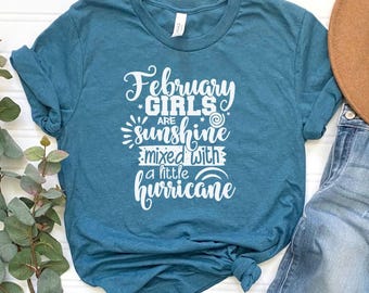 May include: Teal-colored t-shirt with the text "February Girls are sunshine mixed with a little hurricane" in white script. The shirt is a crew neck style and has rolled-up sleeves. A brown hat and blue jeans are partially visible.