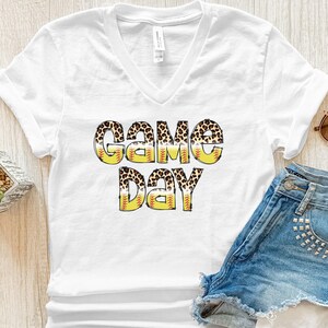 May include: White V-neck t-shirt with the words "GAME DAY" in a playful design. The letters have a leopard print pattern and are outlined with yellow and red baseball stitching, creating a sporty look. The shirt is paired with denim shorts.