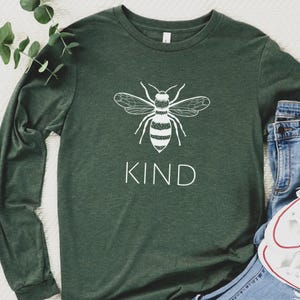May include: A dark green long-sleeved shirt with a white bee graphic and the word "KIND" underneath. The shirt is made of a soft fabric. The design is simple and eye-catching. The shirt is suitable for casual wear.