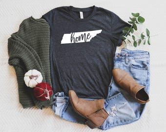May include: A charcoal gray t-shirt with the word "home" in white, shaped like the state of Tennessee. The shirt is paired with blue jeans, brown boots, and a green sweater. Decorative pumpkins are also included.