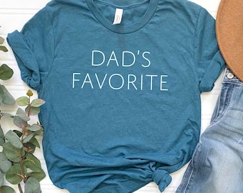 May include: Teal-colored t-shirt with the text "DAD'S FAVORITE" in white. The shirt is made of a soft material and has rolled-up sleeves. The shirt is styled with a brown hat and blue jeans.