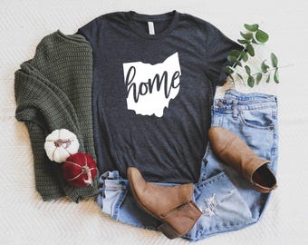 May include: A dark gray t-shirt with the word "home" in a white Ohio outline. The shirt is paired with blue jeans, brown boots, and a green sweater. Decorative pumpkins are also included in the image.