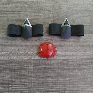 May include: Three-piece set of accessories. Two black elastic bands with gray geometric accents and a red, segmented, shield-shaped piece. The items are arranged on a gray wood-grain surface.