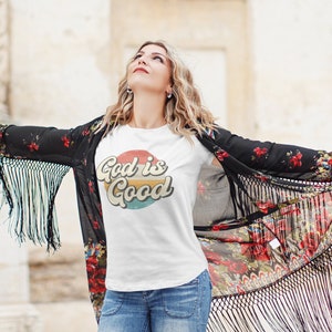 May include: A white t-shirt with a retro-style graphic that says "God is Good" in a colorful circle. The shirt is being worn by a person with long blonde hair and a floral kimono.