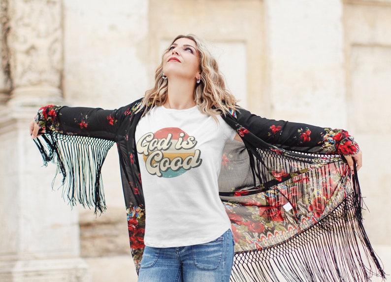 May include: A white t-shirt with a retro-style graphic that says "God is Good" in a colorful circle. The shirt is being worn by a person with long blonde hair and a floral kimono.