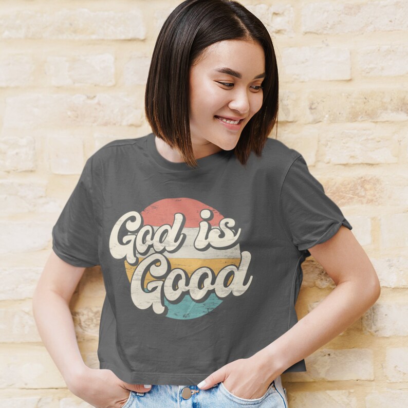 May include: A charcoal grey cropped t-shirt with a vintage-style graphic that says "God is Good" in a colorful circle.