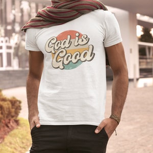 May include: A white t-shirt with a retro-style graphic design featuring a circle with the text "God is Good" in a colorful, layered font.