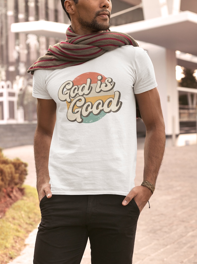 May include: A white t-shirt with a retro-style graphic design featuring a circle with the text "God is Good" in a colorful, layered font.