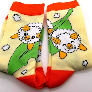 May include: Novelty socks with a cartoon sheep design. The socks have a light yellow background with a green leaf, white clouds, and an orange cuff and toe. The sheep has white fur, orange horns, and a smiling face.