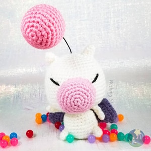 May include: A white crocheted plushie with pink and purple accents. The plushie has a pink ball on top of its head and is surrounded by colourful beads.