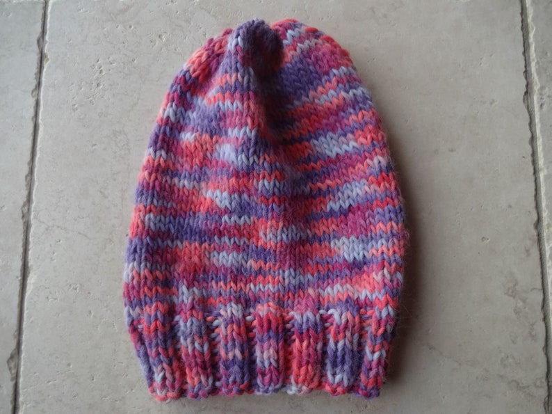May include: A knitted hat with a striped pattern in shades of pink, purple, and white.
