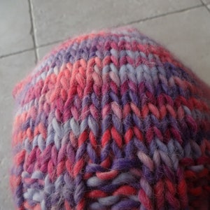 May include: A close-up of a knitted hat in a pink, purple and white colour scheme. The hat is made from a soft, fuzzy yarn.