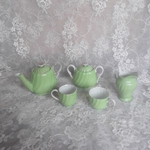 May include: A light green ceramic tea set with a striped design. The set includes a teapot, a creamer, two teacups, and a sugar bowl. The tea set is displayed on a white lace tablecloth.