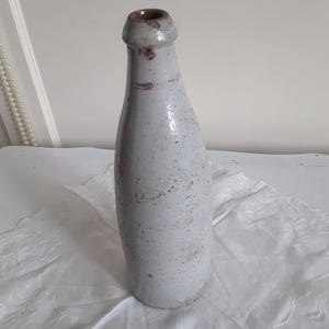 Op de afbeelding: Een vintage, gebroken witte fles met een smalle hals en een licht uitlopende basis. De fles heeft een rustieke, verouderde uitstraling met subtiele bruine spikkels en een donkerbruine opening. Gemaakt van keramiek of steengoed.