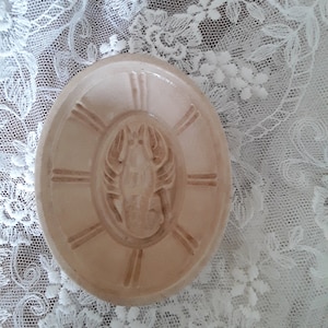 May include: A light peach-colored, oval-shaped ceramic soap mold with a detailed lobster design in the center. The mold has vertical and horizontal lines radiating from the lobster. The mold is set on a white lace background.