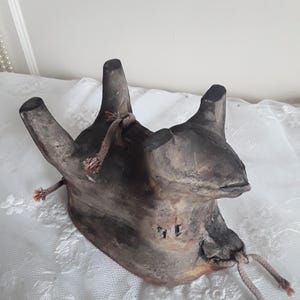 Könnte beinhalten: Dunkelbraunes, handgefertigtes Keramikobjekt mit drei nach oben gerichteten, zylindrischen Beinen. Das Objekt hat eine strukturierte Oberfläche und ist mit einem hellbraunen Seil gebunden. Die Gesamtform ist organisch und rustikal.