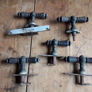 Könnte beinhalten: Sammlung antiker Türgriffe mit dunkler, gealterter Oberfläche. Die Griffe haben ein T-förmiges Design mit dekorativen Details und sind auf verschiedenen Metallplatten montiert. Die Hardware wird auf einer verwitterten Holzoberfläche präsentiert.