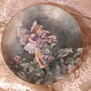 May include: A porcelain plate with a gold rim features a painting of a fairy with wings holding a bouquet of purple flowers. The fairy is wearing a white dress and has a crown of flowers in her hair. The background is a watercolour wash of blue and green.