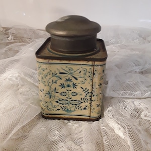 Könnte beinhalten: Eine quadratische Vintage-Blechdose mit einem floralen Design in Blau und Weiß. Der Deckel ist aus Metall und hat eine leicht gewölbte Form.