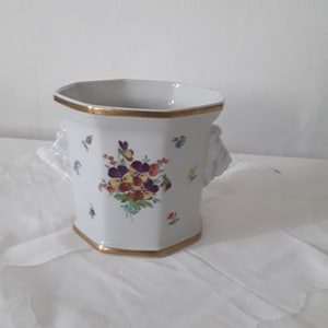 Puede incluir: Jarrón de porcelana blanca con forma hexagonal y ribete dorado. El jarrón presenta un diseño floral con flores moradas, amarillas y naranjas. El jarrón tiene dos asas con cabeza de león.