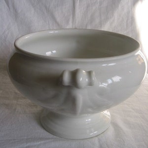 Puede incluir: Un bol de servir de porcelana blanca con un borde acanalado y un asa decorativa. El bol est&aacute; sobre una superficie blanca.