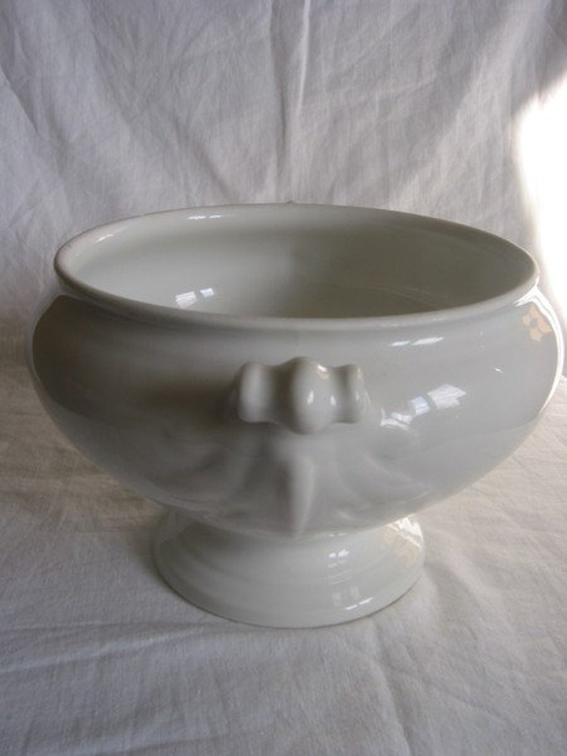 Puede incluir: Un bol de servir de porcelana blanca con un borde acanalado y un asa decorativa. El bol est&aacute; sobre una superficie blanca.