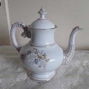 Può includere: Teiera in porcellana bianca con un motivo floreale blu, verde e oro. La teiera presenta un manico e un beccuccio decorativi, con finiture dorate. Il coperchio è sormontato da un terminale. Un pezzo classico per servire il tè.