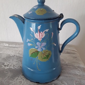 May include: A vintage blue enamel coffee pot with a lid and handle. The pot features hand-painted floral designs in pink, white, and green. The pot is displayed on a white lace tablecloth.