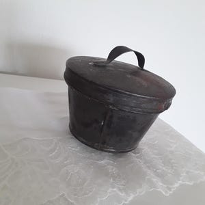 Könnte beinhalten: Ein dunkler Metalleimer mit Deckel und Griff, vermutlich aus der Vintage-Ära. Der Eimer hat eine konische Form und steht auf einem weißen Spitzentuch. Das Metall weist Gebrauchsspuren auf. Der Griff ist einfach und gebogen. Höhe ca. 15 cm.