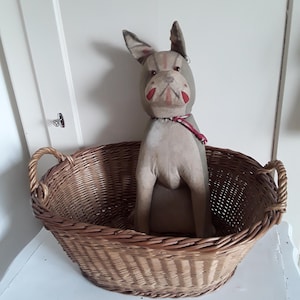 May include: A vintage stuffed dog with a gray body, red accents, and floppy ears sits in a woven wicker basket. The dog has a red collar and is positioned inside the oval-shaped basket, which has handles.